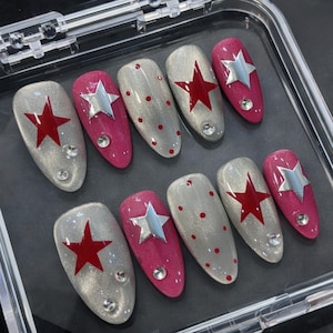 May include: A set of ten artificial nails in a clear plastic case. The nails feature a variety of designs, including silver glitter, red stars, silver stars, red dots, and rhinestones. The nails are oval-shaped and in shades of silver and pink.