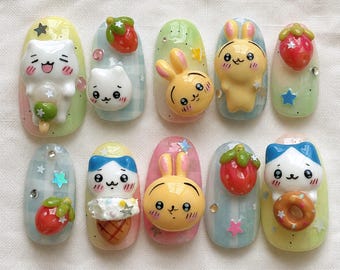 May include: A set of ten press-on nails featuring 3D charms of white cats, yellow rabbits, and red strawberries. The nail bases are decorated with pastel gradients, blue and white gingham patterns, and small star-shaped sequins and rhinestones.