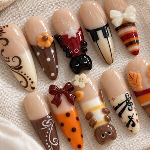 May include: A set of ten almond-shaped press-on nails featuring a nude base with various decorative elements including brown swirls, orange polka dots, a 3D ladybug, a 3D bee, a white bow, a brown bow, piano keys, musical notes, and autumn leaves.
