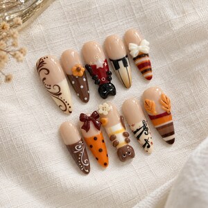 May include: A set of ten almond-shaped press-on nails featuring a nude base with various decorative elements including brown swirls, orange polka dots, a black cat, piano keys, a white bow, musical notes, autumn leaves, and striped patterns.
