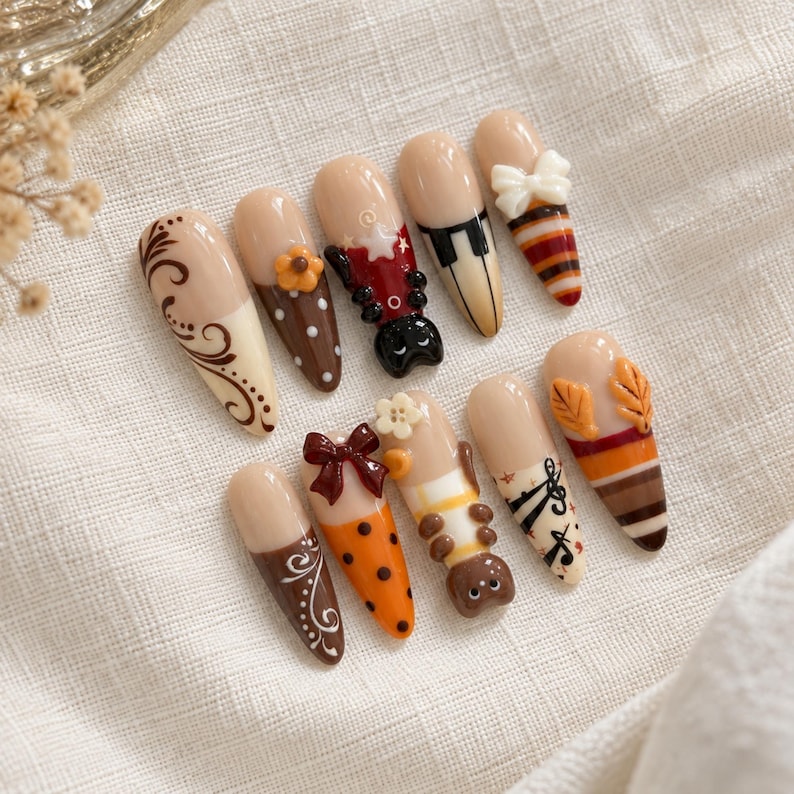 May include: A set of ten almond-shaped press-on nails featuring a nude base with various decorative elements including brown swirls, orange polka dots, a black cat, piano keys, a white bow, musical notes, autumn leaves, and striped patterns.
