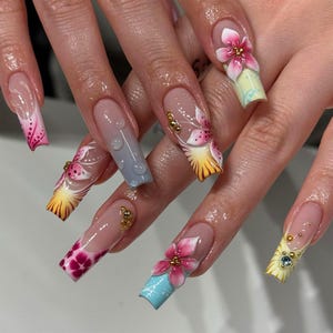 May include: Long, square-shaped acrylic nails with a clear base and intricate floral designs. Each nail features a different design, including 3D flowers, gradients, and gold accents. The color palette includes pink, yellow, blue, and clear.