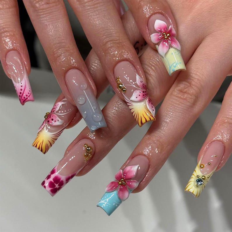 May include: Long, square-shaped acrylic nails with a clear base and intricate floral designs. Each nail features a different design, including 3D flowers, gradients, and gold accents. The color palette includes pink, yellow, blue, and clear.