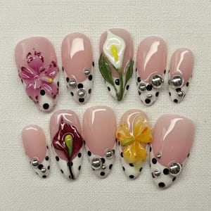 Puede incluir: Juego de diez uñas artificiales con base rosa claro, puntas de lunares blancos y diseños florales en 3D. Algunas uñas presentan adornos plateados. Las flores son en tonos rosa, amarillo y blanco.