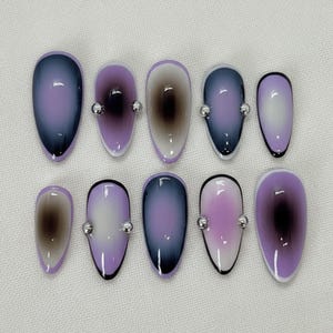 Puede incluir: Un conjunto de diez uñas postizas de forma ovalada. Cada uña presenta un diseño degradado en tonos morados, grises y marrones, con un pequeño adorno de cuentas plateadas. Las uñas tienen un acabado brillante y una base blanca.