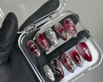 Puede incluir: Un juego de diez uñas artificiales en una caja de plástico transparente. Las uñas son de forma ovalada con un diseño rojo oscuro y plateado. Algunas uñas tienen adornos plateados, otras un efecto degradado.
