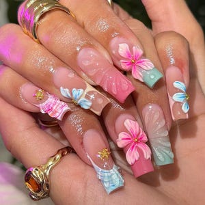 Könnte beinhalten: Nahaufnahme von langen, geformten Fingernägeln mit verschiedenen Designs. Einige Nägel weisen 3D-Blumen in Rosa und Blau mit goldenen Akzenten auf. Andere haben rosa, blaue und weiße Muster. Die Nägel sind in einer transparenten, rosa und nudefarbenen Farbpalette lackiert.