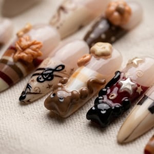 May include: A set of press-on nails featuring various autumn-themed designs including 3D sculpted elements like a brown leaf, a pumpkin, a small brown animal, and a star. The nails display patterns of stripes, musical notes, and solid colors.