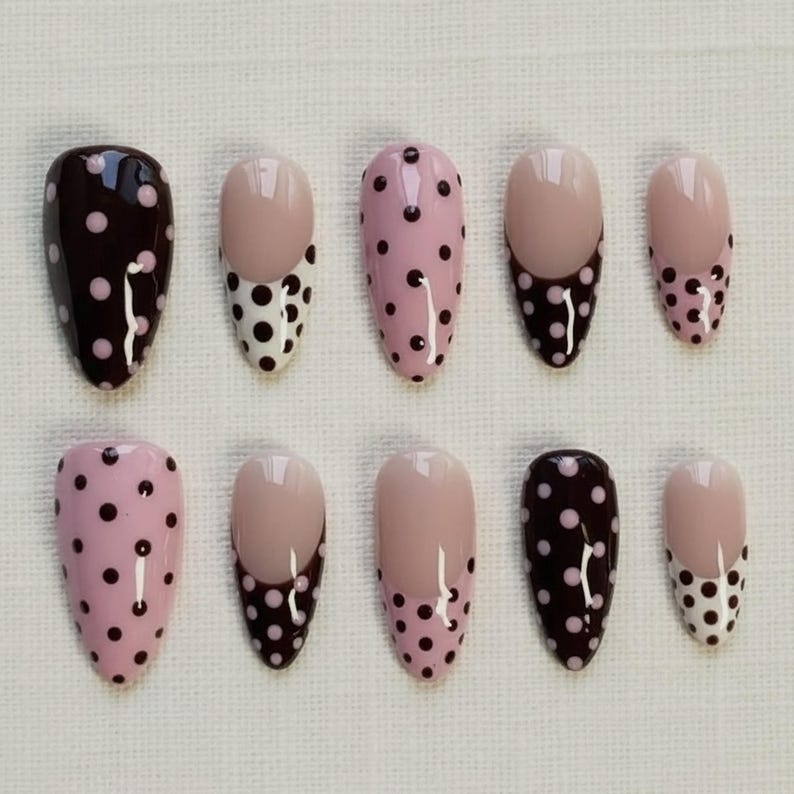May include: A set of ten oval-shaped false nails. The nails feature various designs, including solid colors, polka dots, and French tips. Colors include dark brown, light pink, and nude. The polka dots are black.