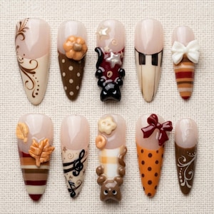 May include: A set of ten press-on nails featuring various autumn-themed designs including brown polka dots, a black cat, a white bow, orange stripes, maple leaves, musical notes, a brown bear, and intricate swirls in shades of brown, orange, and cream.