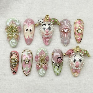 Op de afbeelding: Een verzameling van tien kunstnagels met diverse ontwerpen in roze, groen en goud. Sommige nagels hebben bloemmotieven, terwijl andere een clowngezicht of een ster tonen. De nagels zijn gerangschikt op een witte ondergrond.