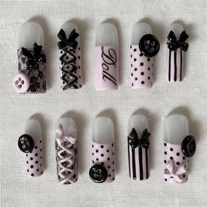 May include: A collection of ten artificial nails with various designs. Some nails feature black bows, lace, polka dots, and vertical stripes. One nail has the word "Doll" written on it, and some have black buttons.