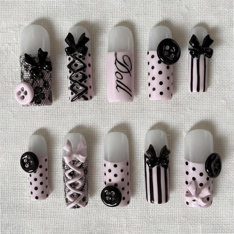 May include: A collection of ten artificial nails with various designs. Some nails feature black bows, lace, polka dots, and vertical stripes. One nail has the word "Doll" written on it, and some have black buttons.