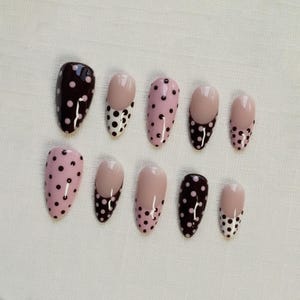 May include: A set of ten artificial nails in various designs. Some nails are solid dark brown with small pink polka dots. Others are light pink with dark brown polka dots. Some have a light pink base with a dark brown polka dot design.