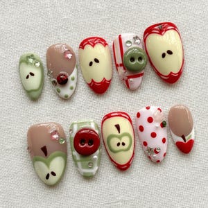 Pode incluir: Uma coleção de dez unhas artificiais com vários designs com tema de maçã. As unhas apresentam uma variedade de cores, incluindo vermelho, verde e bege, com detalhes como botões, bolinhas e enfeites de pedras preciosas. As unhas estão dispostas em uma superfície branca.