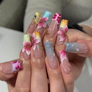 May include: A set of long, acrylic nails with intricate designs. Each nail features a different design, including 3D floral accents, gradient colors, and detailed patterns. The nails are adorned with gold embellishments and water droplet effects.