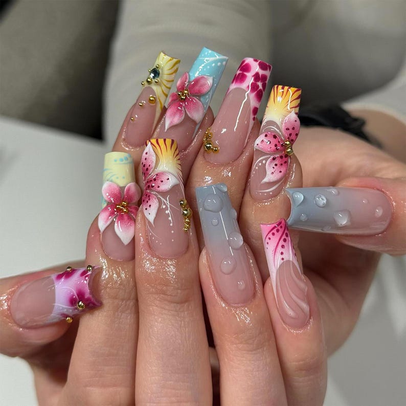 May include: A set of long, acrylic nails with intricate designs. Each nail features a different design, including 3D floral accents, gradient colors, and detailed patterns. The nails are adorned with gold embellishments and water droplet effects.