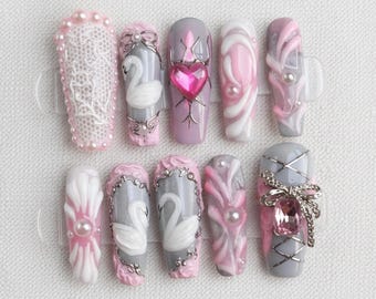 May include: A collection of ten artificial nails with various designs. The nails feature a range of colors, including pink, white, and gray, and are adorned with embellishments such as pearls, lace, and swan motifs. Some nails have heart and bow accents.