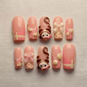 Op de afbeelding: Een set van tien kunstnagels in verschillende ontwerpen. De nagels zijn voornamelijk roze met accenten van wit, bruin en geel. Sommige nagels hebben 3D-versieringen, waaronder strikken, bananen en een apengezicht.