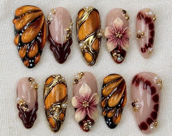 May include: A set of ten press-on nails featuring a mix of designs including metallic gold and copper teardrop patterns, deep burgundy accents, and three-dimensional floral motifs, all embellished with small gold and white spherical beads.