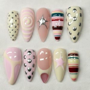 May include: A collection of ten artificial nails in various designs. The nails feature a range of colors, including pink, cream, and silver. Some nails have a swirl pattern, while others have stripes, star accents, or silver studs. The nails are almond-shaped.