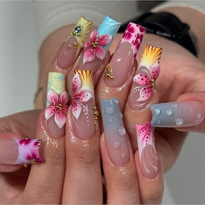 May include: A set of long, artificial nails with intricate floral designs. Each nail features a different design, including pink and white flowers, gold accents, and a gradient effect. Some nails have water droplet details.