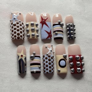 May include: A collection of ten artificial nails with various designs. Some nails feature brown polka dots, silver studs, or star accents. Others display stripes in pastel colors or a red star design. The nail tips are a natural pink.