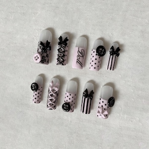 May include: A set of ten artificial nails with various designs. The nails feature black bows, lace, polka dots, and the word "Doll" on one nail. Some nails have button embellishments. The color palette includes pink, black, and white.