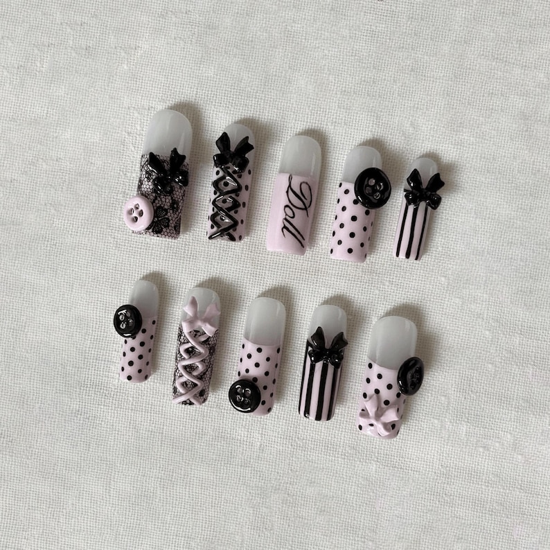 May include: A set of ten artificial nails with various designs. The nails feature black bows, lace, polka dots, and the word "Doll" on one nail. Some nails have button embellishments. The color palette includes pink, black, and white.