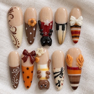 May include: A set of ten almond-shaped press-on nails featuring various designs including brown swirls, polka dots, a black cat, piano keys, a white bow, a dark red bow, a white flower, a brown bear, musical notes, and autumn leaves.