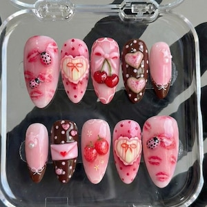 May include: A set of ten artificial nails with various designs. The nails feature pink and brown bases with designs including ladybugs, lips, hearts, bows, cherries, and strawberries. Some nails have a glossy finish, while others have a matte appearance.
