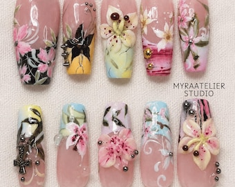 May include: A set of ten press-on nails featuring a nude base with intricate hand-painted floral designs, 3D flower embellishments, metallic studs, and black cross accents. The text MYRAATELIER STUDIO is printed in black on the right side.
