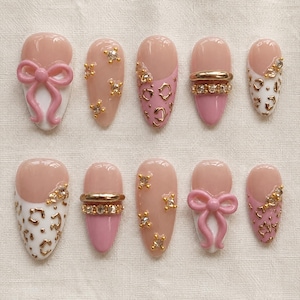 Peut inclure: Un ensemble de dix faux ongles avec une base nude et divers éléments décoratifs, notamment des nœuds roses en 3D, des motifs léopard dorés, des strass transparents et des bandes métalliques dorées, disposés en une collection coordonnée.