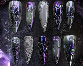 May include: A collection of ten long, pointed artificial nails. Each nail features a unique design with iridescent colors, including purple, green, and silver. Some nails have intricate, web-like patterns, while others showcase swirling textures and embedded gems.