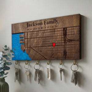 Può includere: Un portachiavi in legno con un design a mappa, con il testo "Jackson Family" e le coordinate. La mappa include una sezione blu e un indicatore rosso. Diverse chiavi sono appese a ganci in ottone sottostanti.