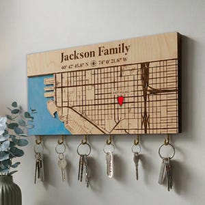 May include: A wooden key holder with a map design, the text "Jackson Family", and coordinates. The map is partially blue, with a red marker. Below the map are several key hooks with keys hanging from them.