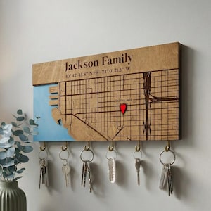 May include: Wooden key holder with a map design, featuring the text "Jackson Family" and coordinates. The map includes a red marker and several key hooks. The key holder is mounted on a wall, with keys hanging from the hooks.