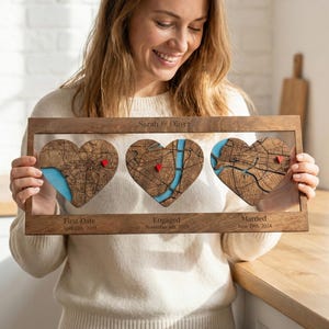 May include: A wooden framed piece of art with three heart-shaped maps, each with a red pin and blue detailing. The text "Sarah & Oliver" is above the hearts. Below, the text reads "First Date," "Engaged," and "Married" with dates.