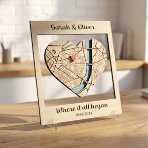 Può includere: Una mappa a forma di cuore in legno con un perno rosso, incorniciata in una cornice di legno chiaro. La cornice ha i nomi "Sarah & Oliver" in alto e la frase "Where it all began" con una data in basso. La mappa mostra una pianta della città.