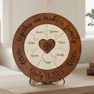 May include: A wooden family puzzle with a dark brown frame and light wood puzzle pieces. The puzzle reads "together we make a family" and includes names and the date "July 17, 2023". A heart-shaped tree design is in the center.