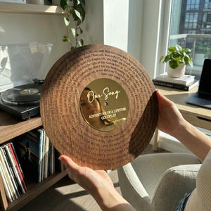 Puede incluir: Un disco de madera con letras grabadas y un centro dorado. El texto "Our Song" y "Adventure of a Lifetime Coldplay" son visibles. El disco se sostiene frente a una persona.