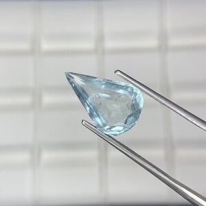 May include: A pear-shaped, light blue, faceted gemstone held by silver-toned metal tweezers. The stone is translucent with visible internal facets and a pointed top, showcasing a clear, icy blue colour throughout its polished, geometric surface.