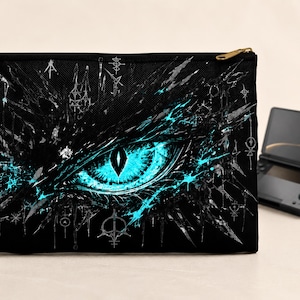 May include: Black zippered pouch featuring a glowing teal dragon eye design. The eye is the focal point, with intricate details and a dark background. A black handheld gaming device is visible in the background.