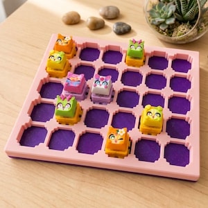 May include: A square pink grid tray with a purple base featuring several small, colourful, character-shaped keycaps placed in the slots. The keycaps include various designs such as orange, green, and patterned figures with distinct facial expressions.