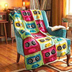 Può includere: Una colorata coperta all'uncinetto drappeggiata su una poltrona turchese. La coperta presenta un motivo ripetuto di musicassette in colori vivaci come rosa, giallo, verde e blu. I disegni delle musicassette hanno dettagli neri e grigi.