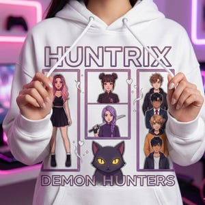 May include: White hoodie with a graphic featuring the text "HUNTRIX DEMON HUNTERS." The design includes anime-style illustrations of characters and a black cat with yellow eyes, all within a purple frame.