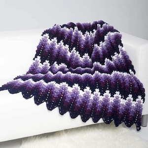 May include: A handmade crochet blanket in shades of purple, white, and dark blue, draped over a white sofa. The blanket features a chevron pattern with a scalloped edge, creating a textured and visually appealing design.