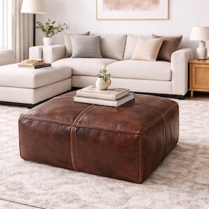 Puede incluir: Un gran puf cuadrado de cuero marrón con costuras decorativas. El puf es el punto focal de un salón, con un sofá blanco y una decoración de tonos neutros en el fondo. Libros y un pequeño jarrón encima.