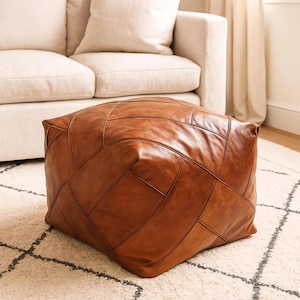Może przedstawiać: Kwadratowy, brązowy puf ze skóry z patchworkowym wzorem. Puf stoi na kremowym dywanie z czarnym geometrycznym wzorem. Beżowa sofa i zasłony są widoczne w tle.