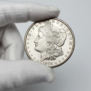Puede incluir: Una moneda de plata con el perfil del rostro de una mujer, rodeada de texto y estrellas. La moneda está fechada en 1878. La moneda está sujeta por una mano enguantada.